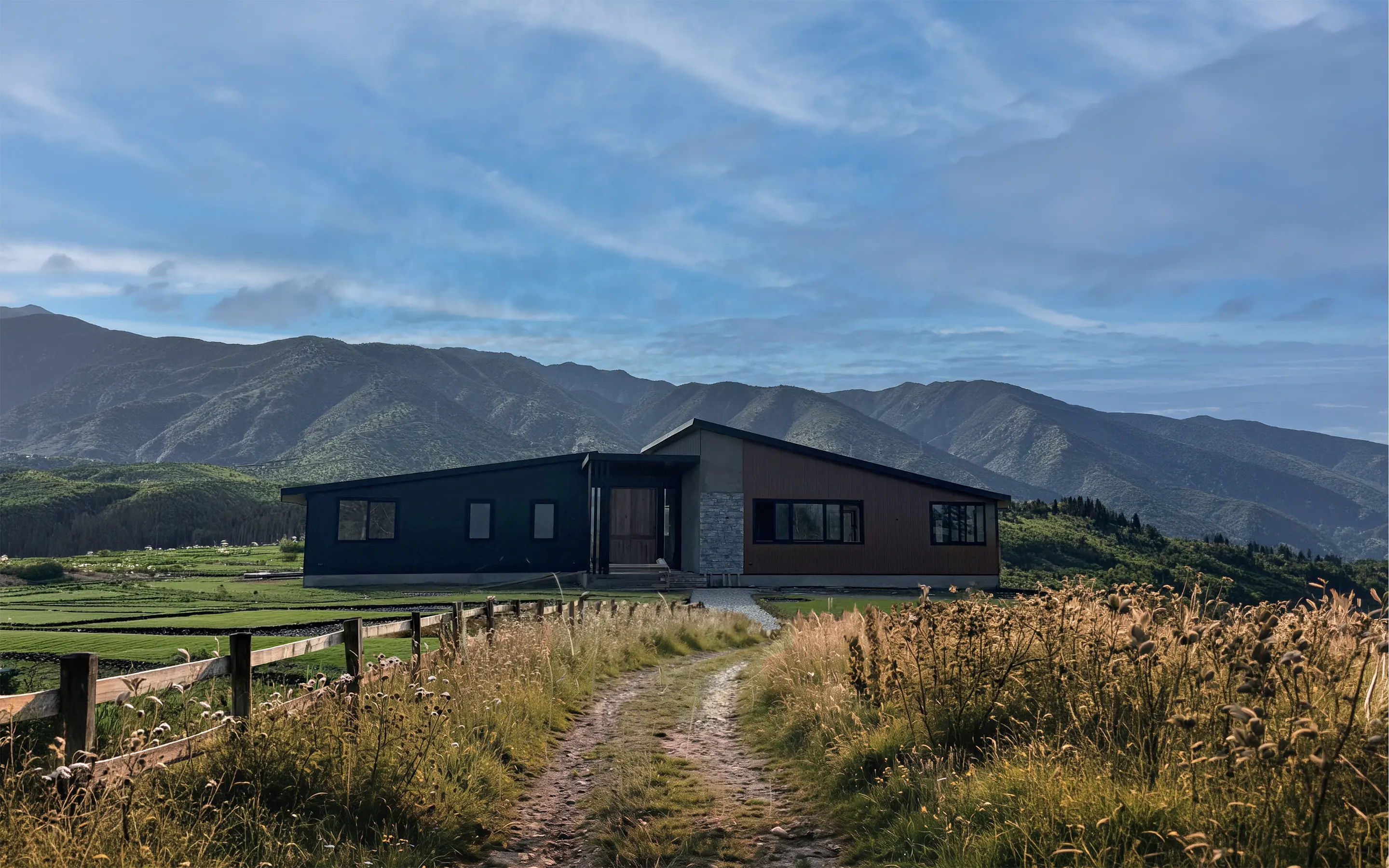 Casa en Santa Luisa de Palpi vista desde el camino, entre pasto alto con espigas y un cerco de madera, con los cerros al fondo